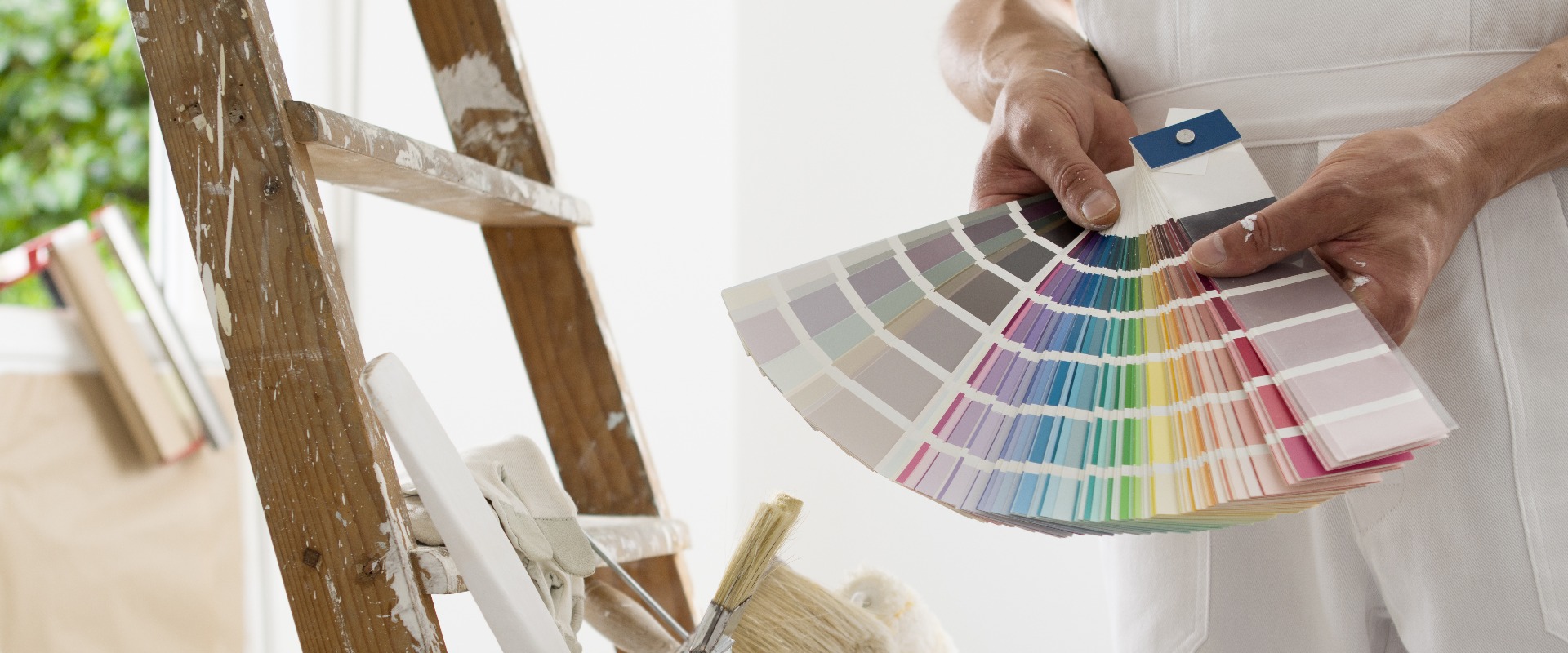 Die Hände eines Malers und Dekorateurs, der anhand eines Farbmusters die Farbe auswählt; im Hintergrund sind die zu renovierenden Räume, eine Holzleiter sowie Pinsel und ein Eimer zu sehen; Nahaufnahme